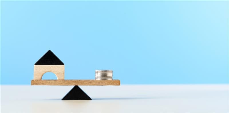 A small wooden house on one side of a scale with a small stack of coins on the other, representing risk weighted assets